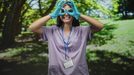 Woman nurse in purple scrubs wearing blue gloves forms finger goggles with hands and sticks out tongue in forest; playful compassion.