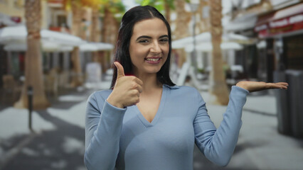 Hispanic woman outdoor giving thumbs up with palm extended in an urban street setting wearing blue...