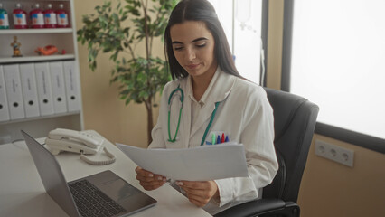 Woman doctor with stethoscope reviews documents in a clinic office, showcasing a professional setting with emphasis on healthcare and focus, surrounded by office essentials.