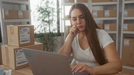 Woman typing on laptop beside stacked parcels processing online orders in office building; pensive small business planning.