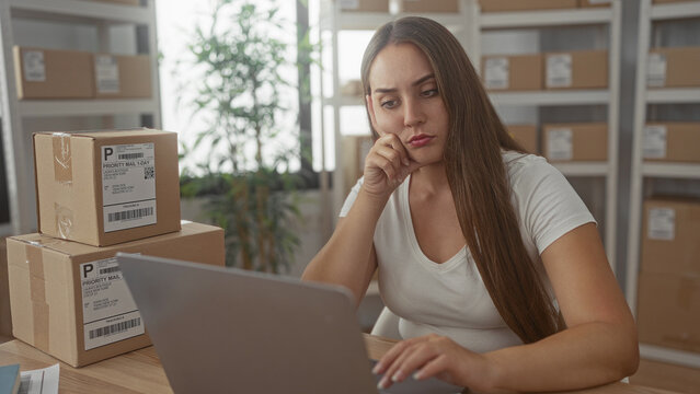Woman typing on laptop beside stacked parcels processing online orders in office building; pensive small business planning.
