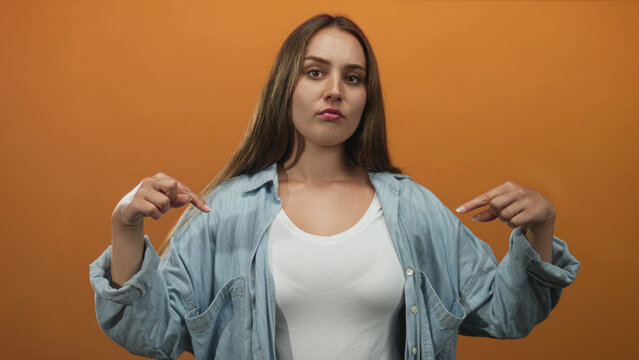 Woman finger pointing to chest wearing white tank top and denim shirt in studio; confidence self esteem.