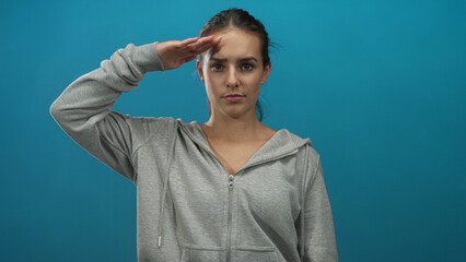 Young hispanic woman in gray hoodie saluting with right hand to forehead in studio teal wall,...