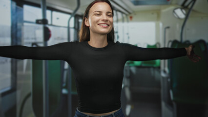 Fototapeta premium Young woman smiling with face turned up and eyes closed, arms fully outstretched across aisle on city bus; joy freedom travel.