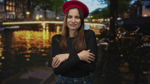 Woman in red beret crosses arms hugging chest on street by amsterdam canal with docked bicycles and lit bridge at night; loneliness.
