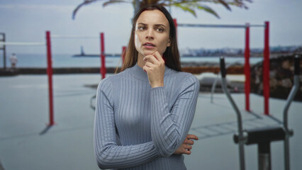 Woman with hand on chin and finger to lips at outdoor gym by the waterfront near red pullup bars and exercise equipment; contemplation doubt.