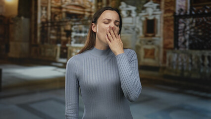 Woman touching head with hand wearing blue ribbed turtleneck sweater in church building interior aisle; quiet sadness reflection.