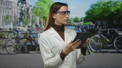 Woman scientist in white lab coat and safety goggles holding tablet on street in amsterdam by canal and bicycles; urban research focus.