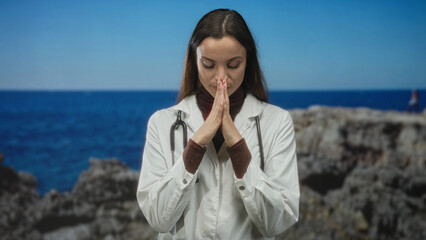 Woman doctor with stethoscope and hands in prayer gesture on coast near rocky shore; compassion...