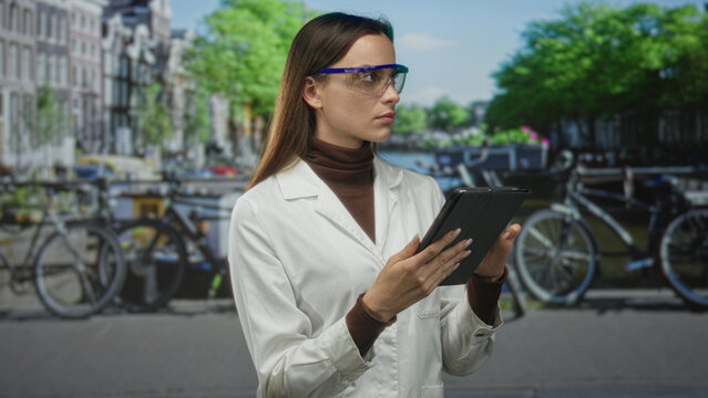 Woman scientist in white lab coat and safety goggles holding tablet on street in amsterdam by canal and bicycles; urban research focus.
