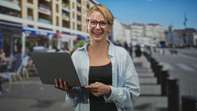 Woman wearing glasses holds laptop with both hands and smiles while typing on a street with cafes and bollards; remote work happy. - Powered by Adobe