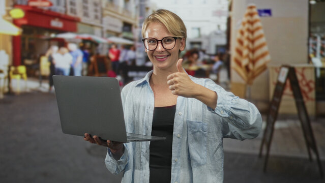 Woman holding laptop and typing with both hands while smiling and wearing glasses on street cafe; remote work content.