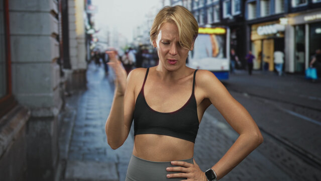 Woman pinches nose with fist near face, sports bra and bare midriff on street with tram visible; disgust.