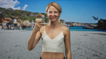Woman holds double scoop ice cream cone in hand on sunny sandy beach, smiling and presenting the treat while glancing at the cone; carefree joy.