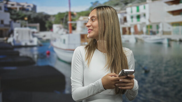 Woman holding smartphone at marina pier, smiling and looking to the side; leisure connection contentment.