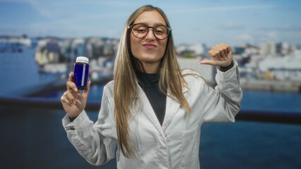 Woman in white lab coat wearing glasses holding a blue bottle and points thumb to chest in a studio...