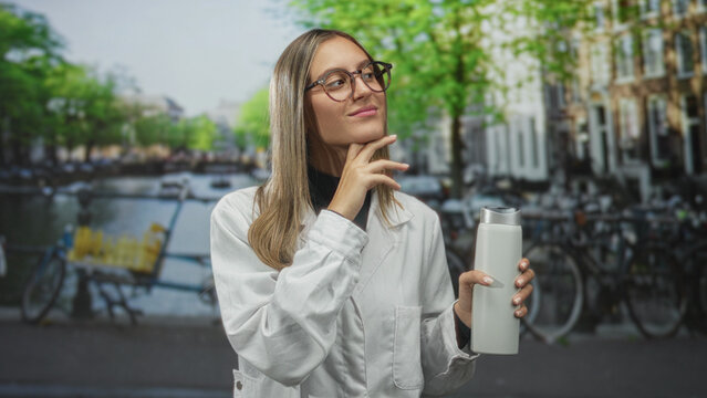 Woman holding a white bottle and touching her chin on an urban street by a canal with parked bicycles and buildings visible; thinking.