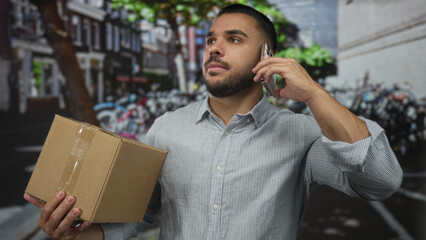 Man holding a cardboard package in one arm and pressing a smartphone to his ear on a city street lined with parked bicycles; urban urgency.