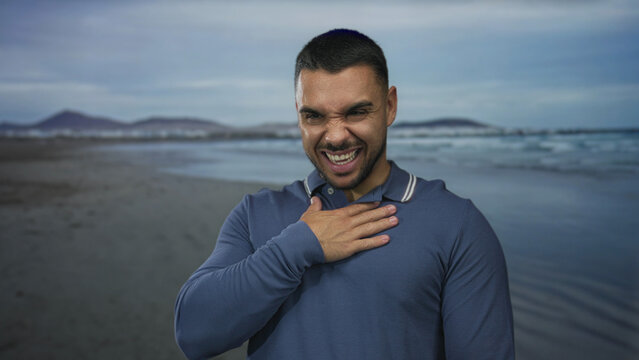 Young hispanic man with hand on chest standing on sandy beach shore by calm ocean water waves; serenity.