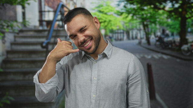 Man wearing striped shirt showing call gesture with hand on street scene; friendly invitation social connection.