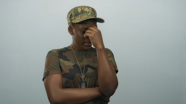 Woman soldier wearing camouflage uniform and dog tags pinches nose in pale blue studio; disgust fatigue distress.