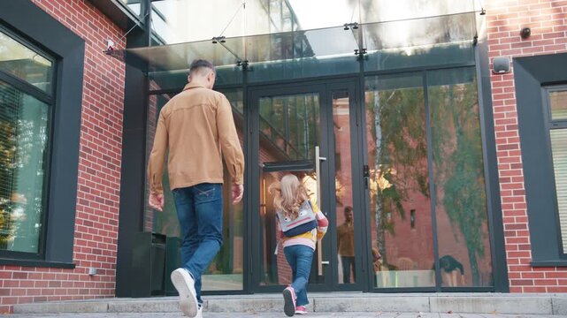 Caucasian father walking beside daughter hopping playfully near school entrance. Adult male watching child with backpack as she moves toward glass doors along brick pathway during lively morning.