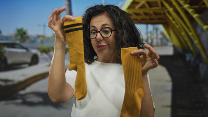 Middle age hispanic woman holds two different socks on city sidewalk; confusion frustration chores.