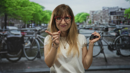 Woman wearing red glasses holding car keys and finger to lips for silence in street in amsterdam; playful secrecy.