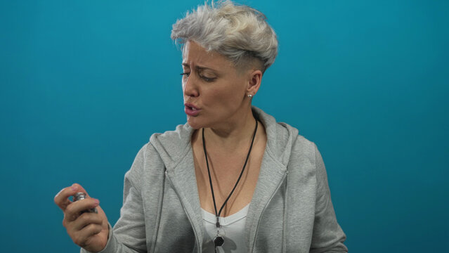Woman checking stopwatch in studio with tense expression, raised hand and anxious glance against a blue backdrop; impatience urgency pressure.