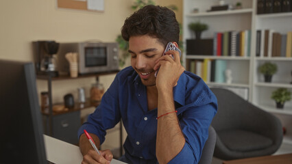 Man in blue shirt holding smartphone to ear and writing with pen at desk in a building; focused productivity.