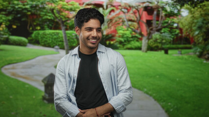 Fototapeta premium Young hispanic man smiling, hands clasped at waist, wearing black tee and open button shirt in a building courtyard; contentment ease.