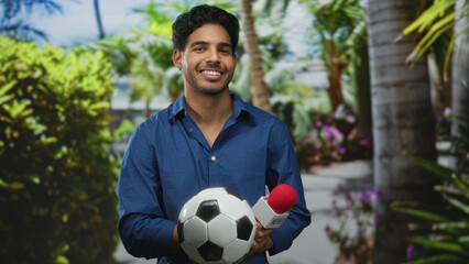 Man holding soccer ball and red foam news microphone, smiling with eyes closed and head tilt in studio; celebration joy.