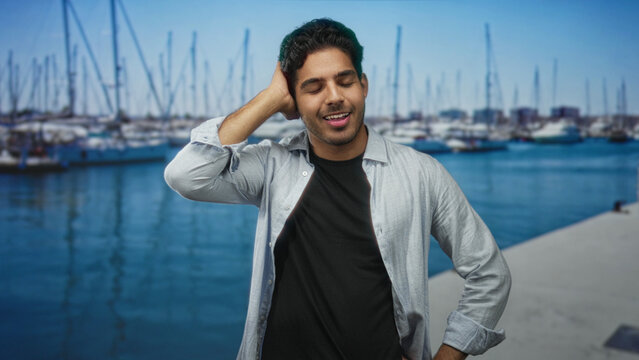 Man in casual shirt touching his head with hand, eyes closed and smiling while standing on a marina street near docked sailboats and water; relaxed contentment.