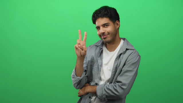 Young hispanic man smiling holds up a peace sign with raised hand against a green screen in studio; friendly optimism.