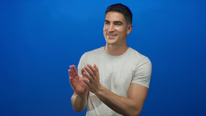 Man clapping hands and smiling in blue studio with white t shirt and casual pose; cheerful appreciation.