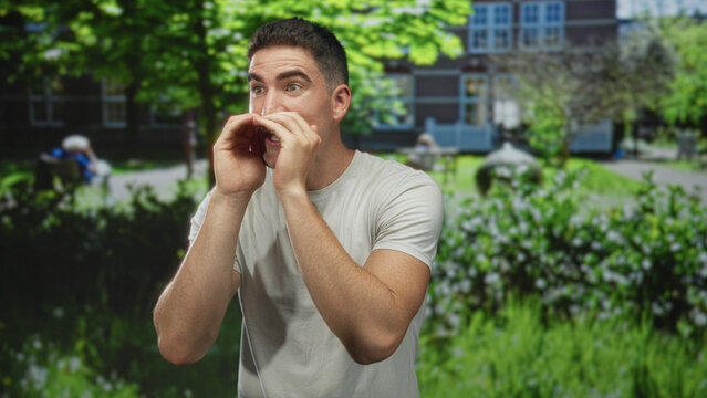 Young hispanic man cups hands to mouth calling across sunlit park lawn near building entrance; confidence outreach.