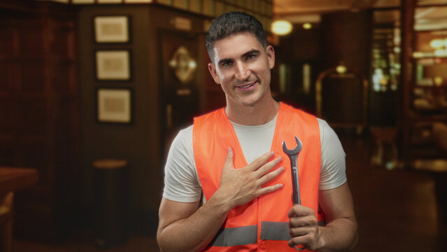 Man engineer wearing orange safety vest holding wrench with hand to chest in hotel building lobby; pride duty. - Powered by Adobe