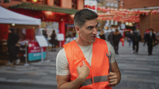 Man technician points finger to chest holding a wrench and wearing orange safety vest on busy street market with lanterns and stalls; pride responsibility.