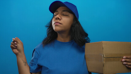 Delivery woman in blue cap and shirt points finger forward and holds cardboard box in studio...