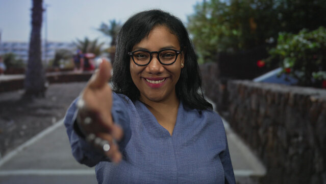 African american woman wearing glasses extends her right hand for a handshake on a city street; friendliness.