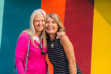 Mature women friends enjoying time together, smiling with arm embrace against a vibrant wall
