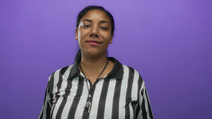 African american woman referee in striped jersey flips hair with hand and smiles in a purple studio setting; confidence empowerment strength spirit.