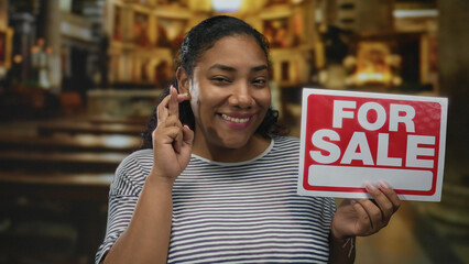Woman holding red for sale sign while crossing her fingers and smiling amid wooden pews and stained...