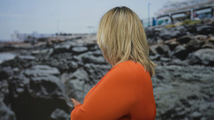 Woman in orange sweater points hand to rocky shore at sunny beach while smiling under clear sky; contentment.