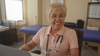Senior physiotherapist woman smiling with stethoscope, visible face and hands, seated by treatment table in a medical building; warmth trust care.