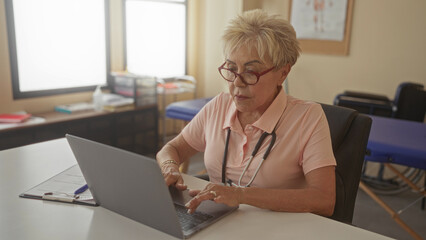 Woman physiotherapist typing on laptop with stethoscope around her neck and reading patient notes at desk in a medical building; calm professional care.