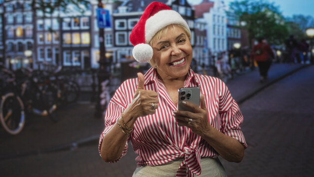 Woman in santa hat holds smartphone and gives thumbs up on a street lined with canal buildings, smiling and checking her phone; holiday joy.