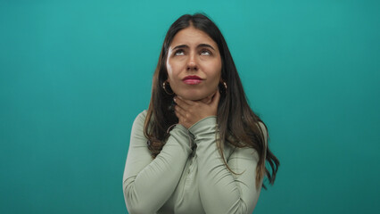 Obraz premium Young hispanic woman holds throat with both hands for a sore throat in a green studio; health concern discomfort.