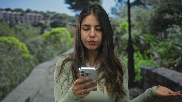 Woman using smartphone in green park on sunny day, showcasing an outdoor lifestyle with focus on technology, young female engaged in digital communication amidst nature. - Powered by Adobe