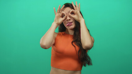 Fototapeta premium Young hispanic smiling woman in orange top makes ok sign binocular gesture in studio; playful curiosity joy.